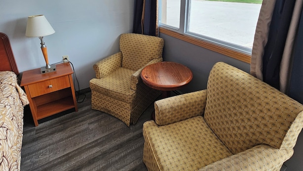 A hotel room with a bed, a nightstand with a lamp, two patterned armchairs, a small wooden table, and a window with a view of the outside. - Forest Plaza Motel (Wellington North)