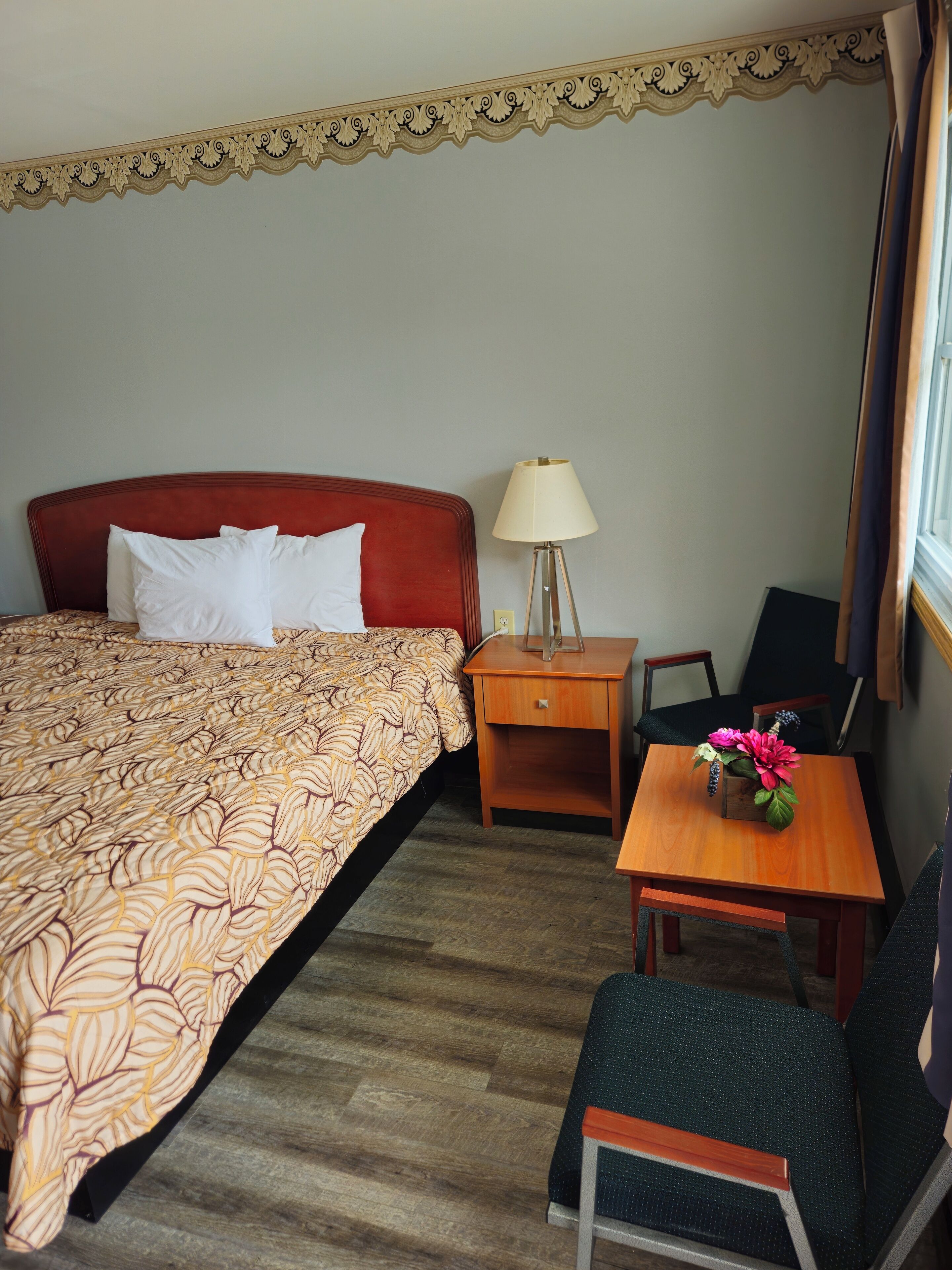 Une chambre d’hôtel avec un lit, une table de chevet, une table ornée d’un vase de fleurs, une chaise et une lampe.