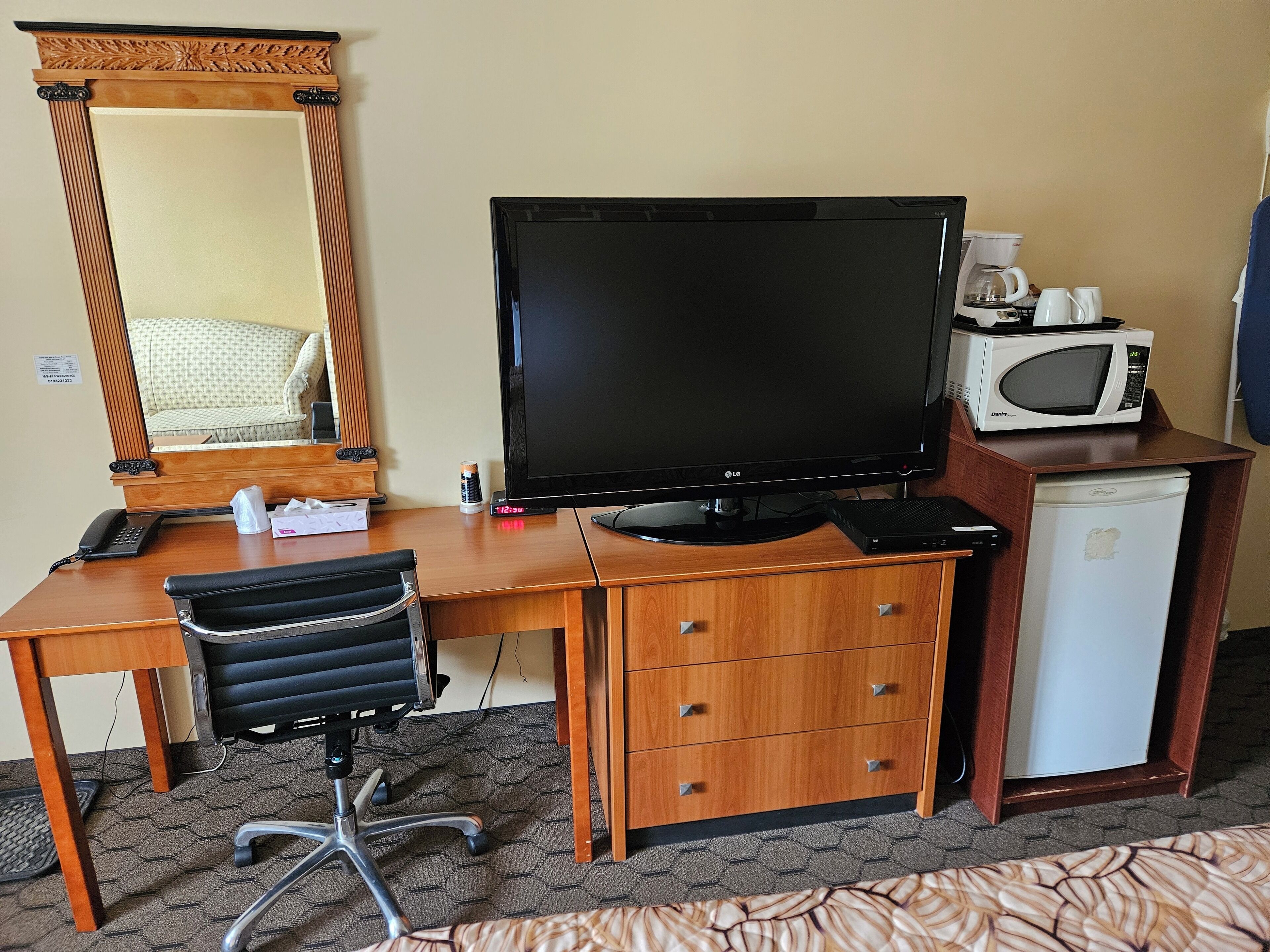 A hotel room with a wooden desk, a large flat-screen TV, a microwave, a small refrigerator, a chair, and a mirror.