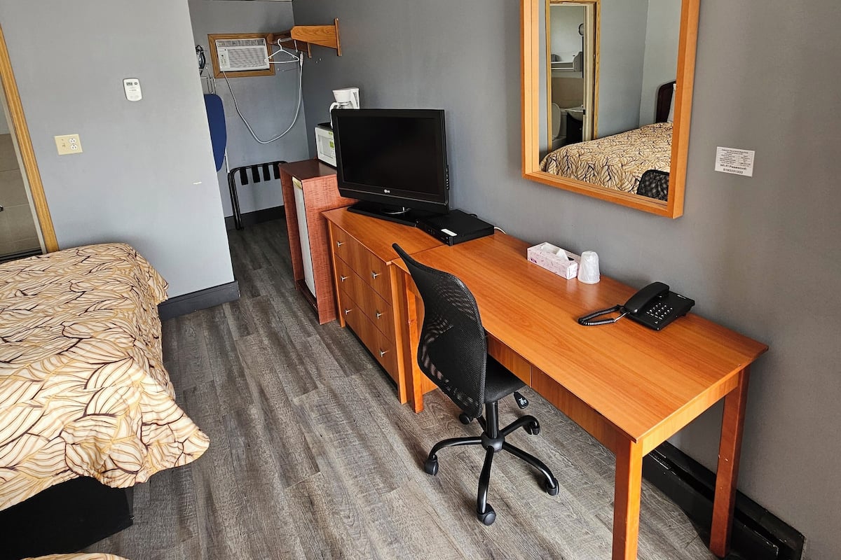 Une chambre d’hôtel compacte comprenant un lit, un bureau avec un ordinateur, une chaise, un miroir et une petite kitchenette.