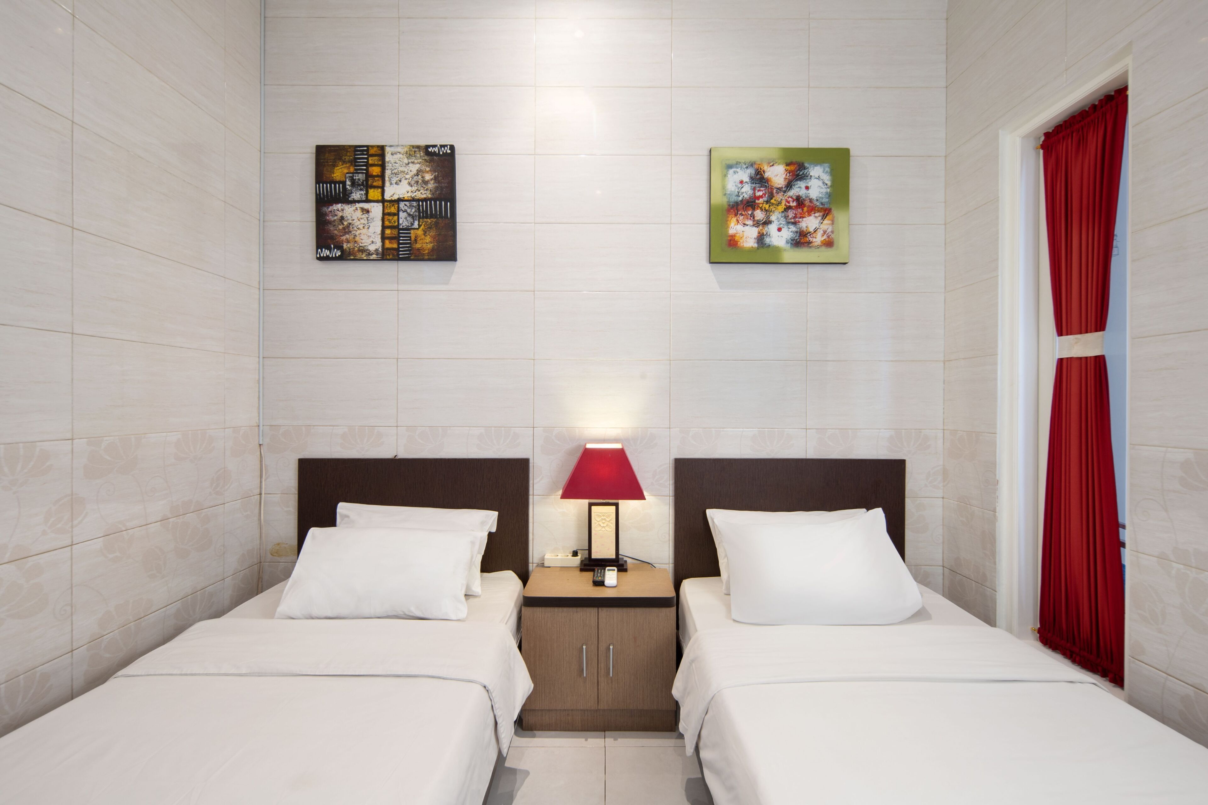 A hotel room with two beds, a nightstand with a lamp, and two artworks on the wall.