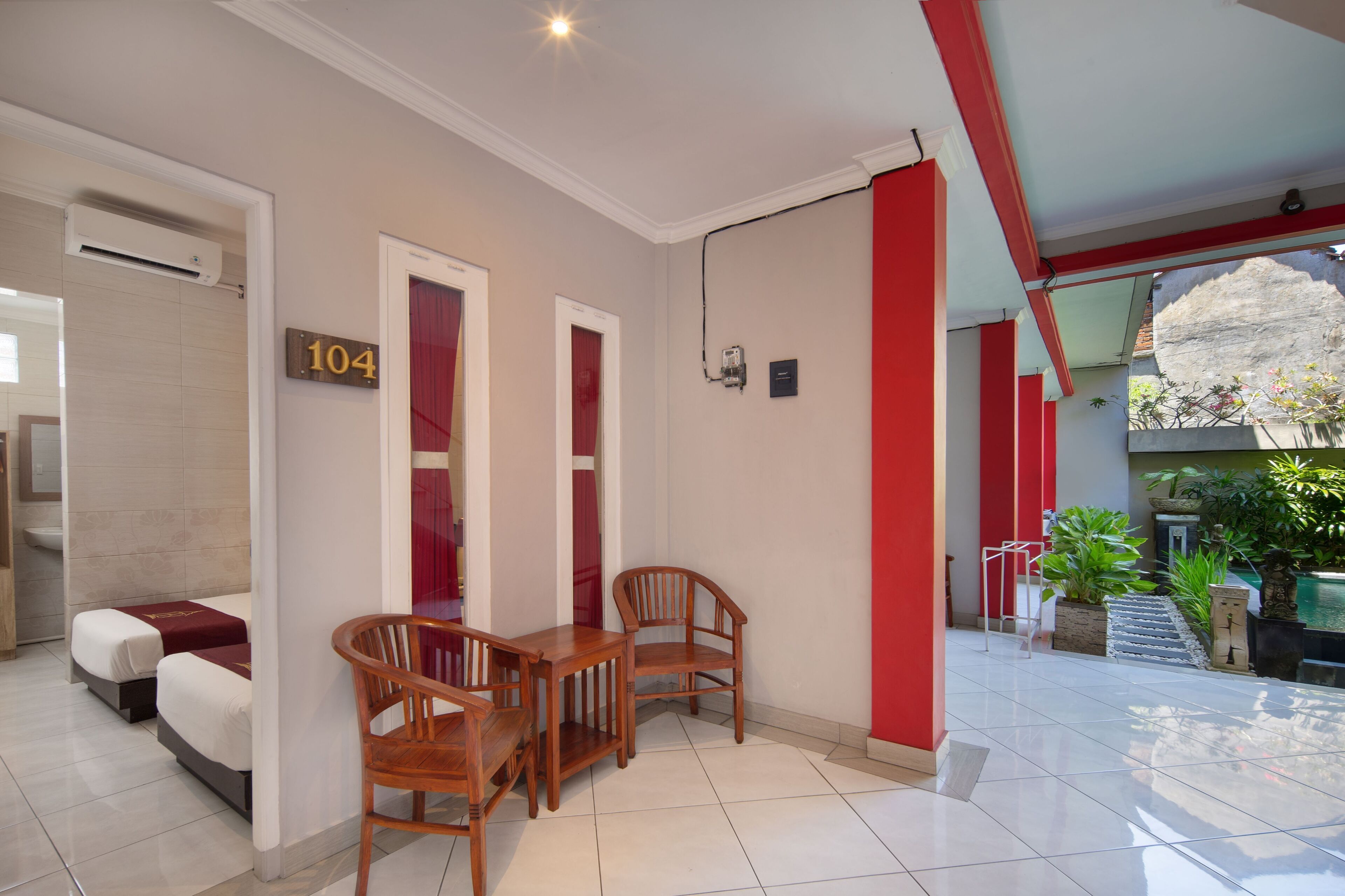 Une chambre d’hôtel avec un lit, une table en bois et deux chaises. Il y a un panneau avec un numéro sur le mur et un pilier rouge.