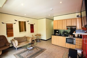 A compact living space with a sofa, a small round table, a kitchen area with wooden cabinets and a microwave, and a hanging light fixture.