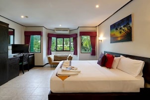A hotel room with a large bed, a desk, and a chair. There are windows with curtains, a painting on the wall, and a television on the desk.