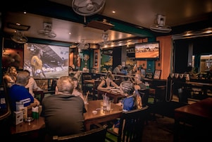 A restaurant with multiple tables, ceiling fans, and a large screen displaying a sports event.