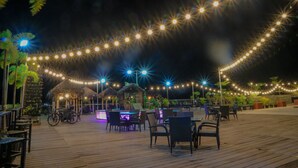 Una terraza de madera con luces cálidas, mobiliario exterior y estructuras de techo de paja.