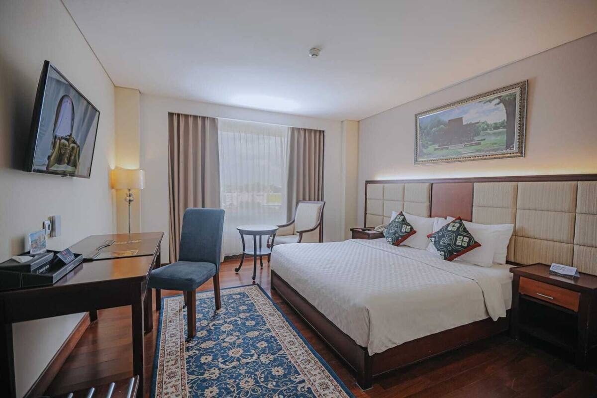 Una habitación de hotel con cama, escritorio, silla, una mesita y un cuadro en la pared.