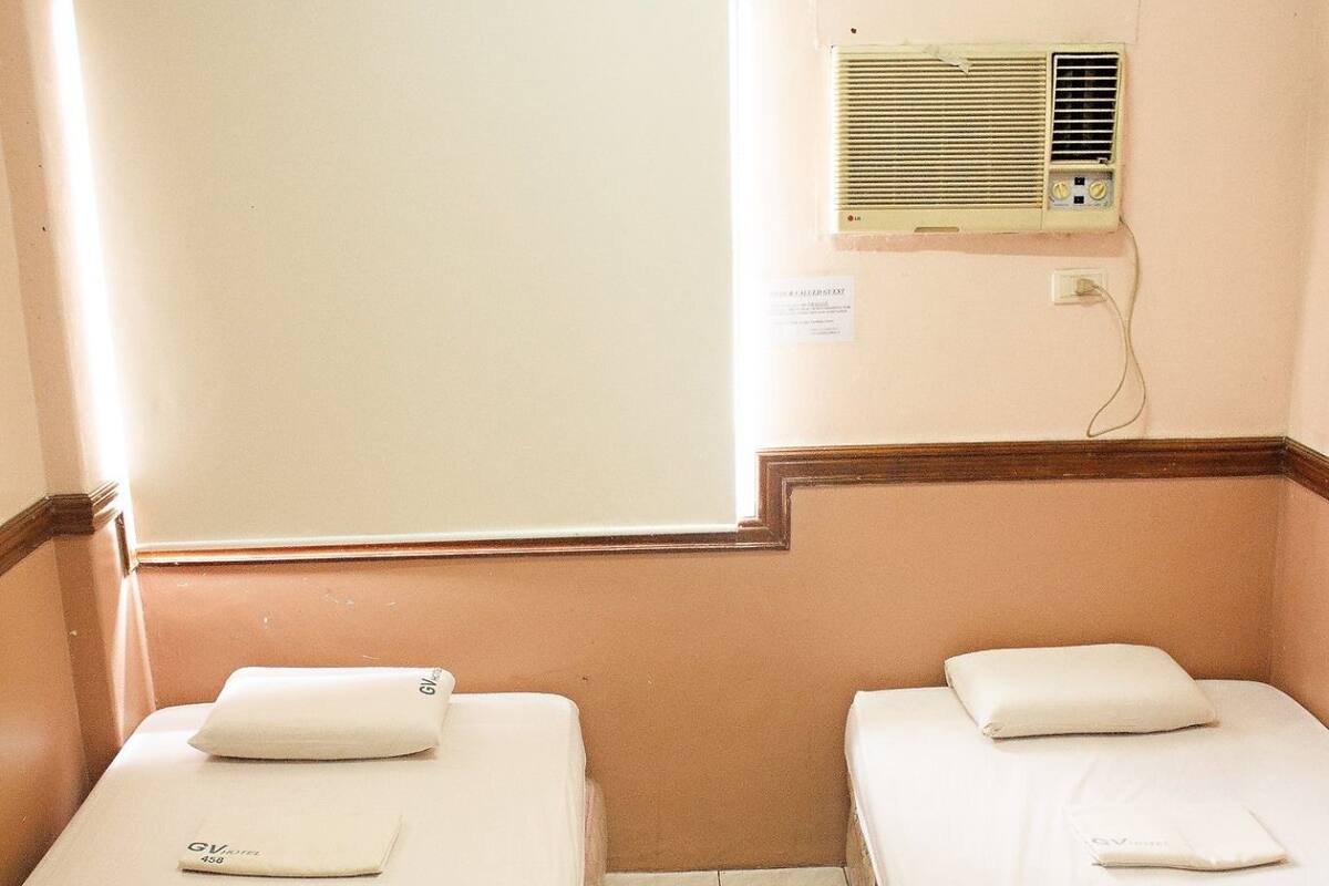 A small hotel room with two single beds, a window, a wall-mounted air conditioner, and a tiled floor.