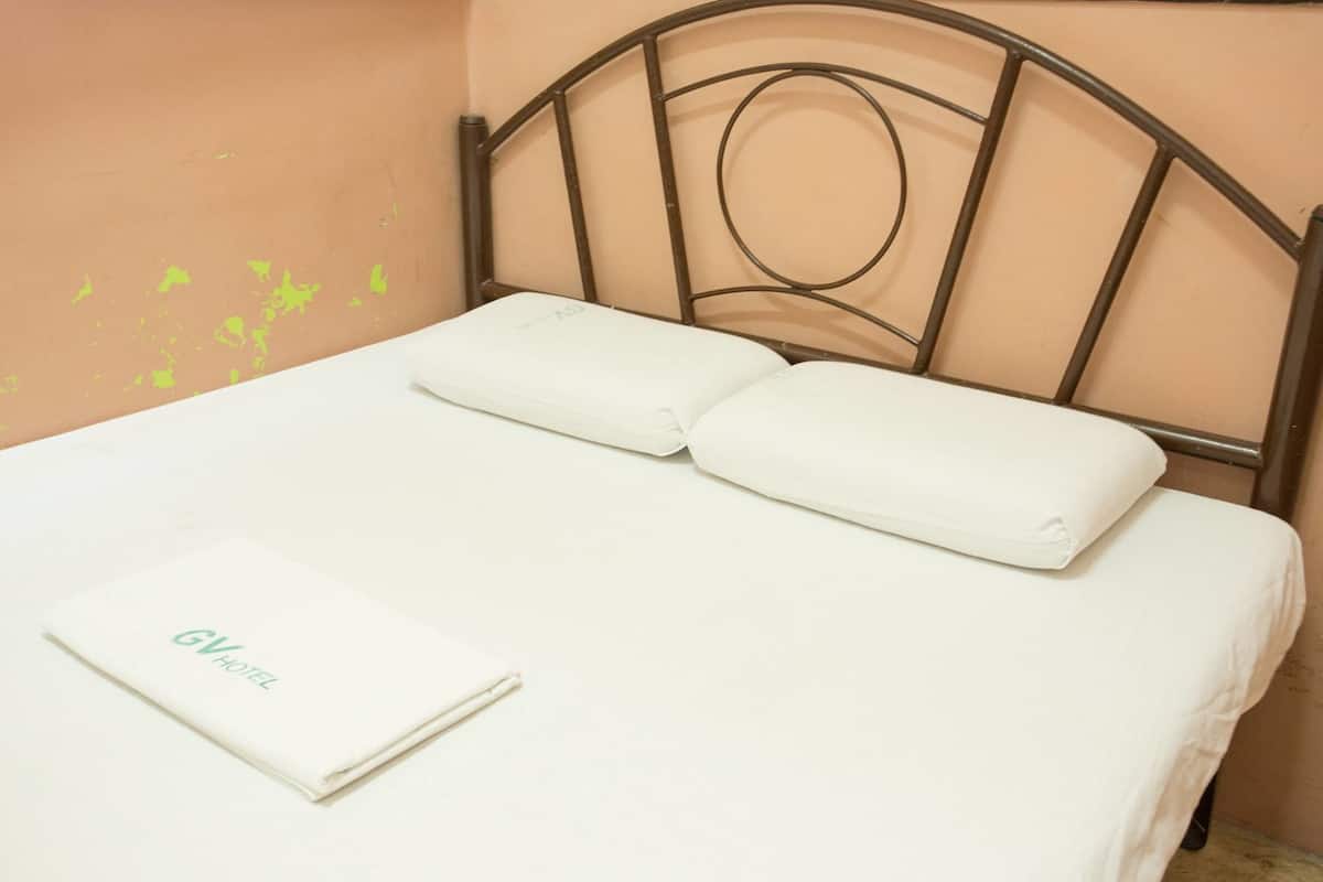 A neatly made bed with white linens and pillows, a folded white towel with "GV Hotel" text, and a metal headboard.