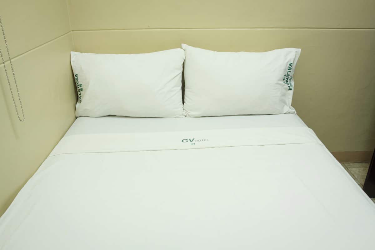 A single bed with white bedding and pillows, a visible logo on the bedspread, and a plain wall with a chain hanging on it.