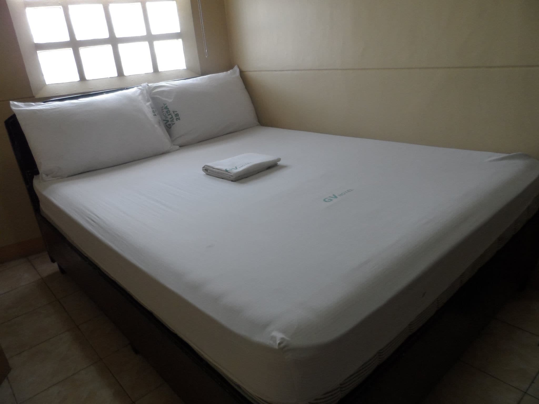 Una cama bien hecha con sábanas blancas y una toalla doblada, una ventana con persianas y una pared lisa.
