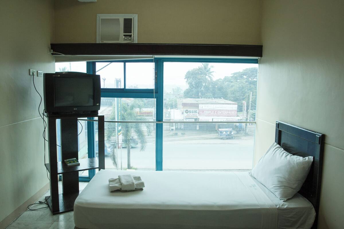 A hotel room with a bed, a television on a stand, and a window with a view of the street and buildings outside.