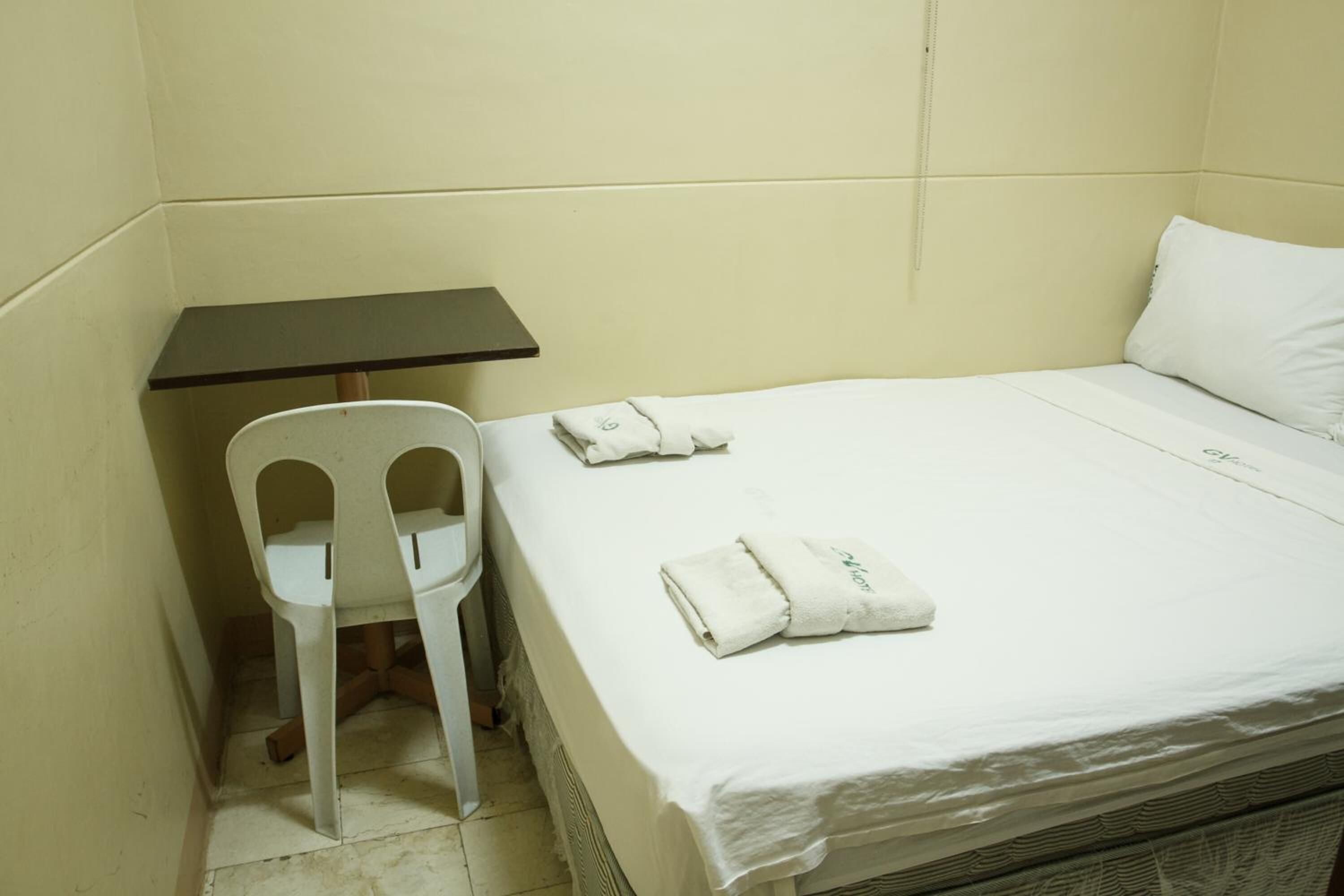 A small, single-bed room with a white bedspread, a white chair, and a small table.