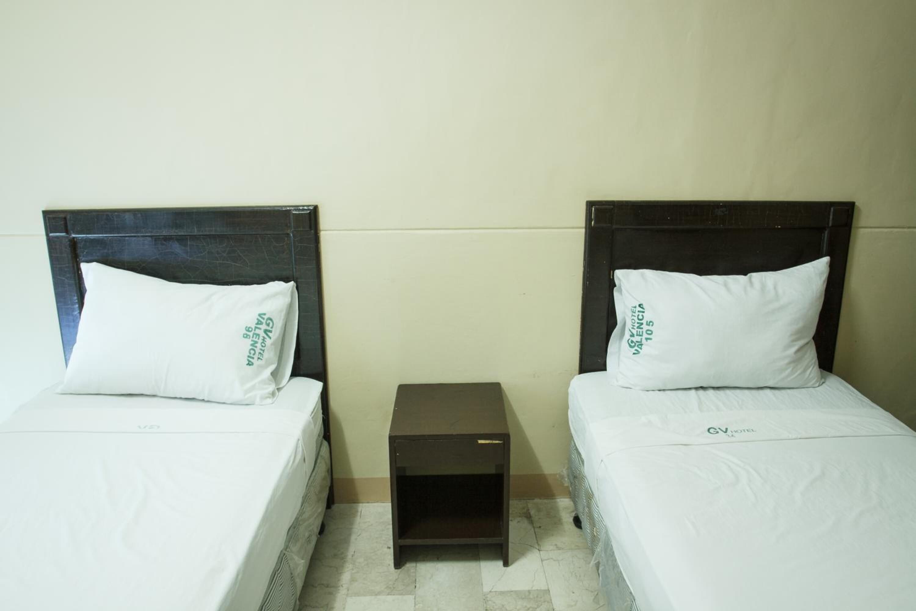 Two single beds with matching headboards and pillows, a small bedside table, and a plain wall.