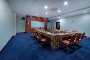 Un salón de conferencias con una larga mesa, sillas rojas y una pantalla de proyector en la pared.