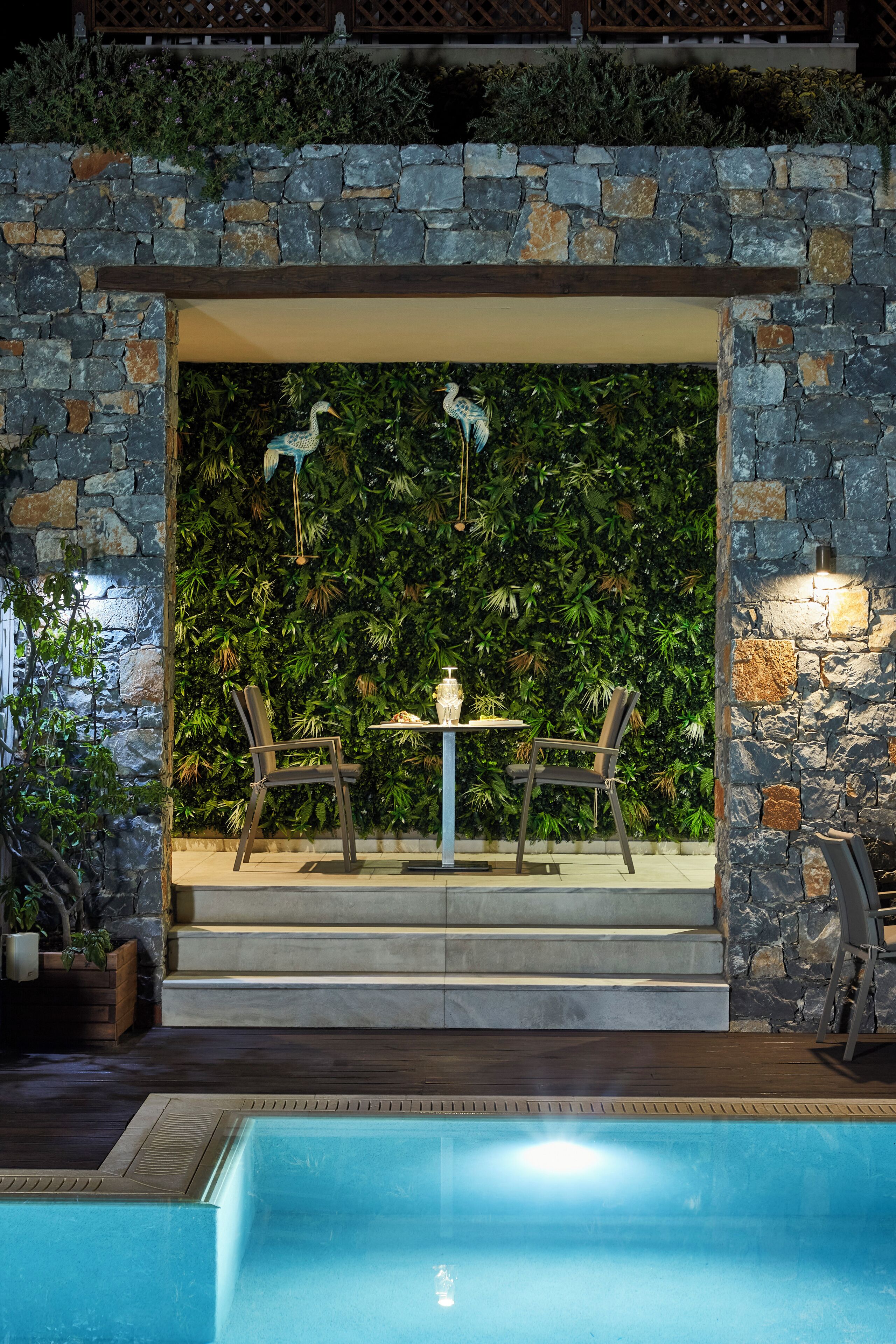 A poolside area with stone walls, two chairs, a table, and a decorative bird sculpture.