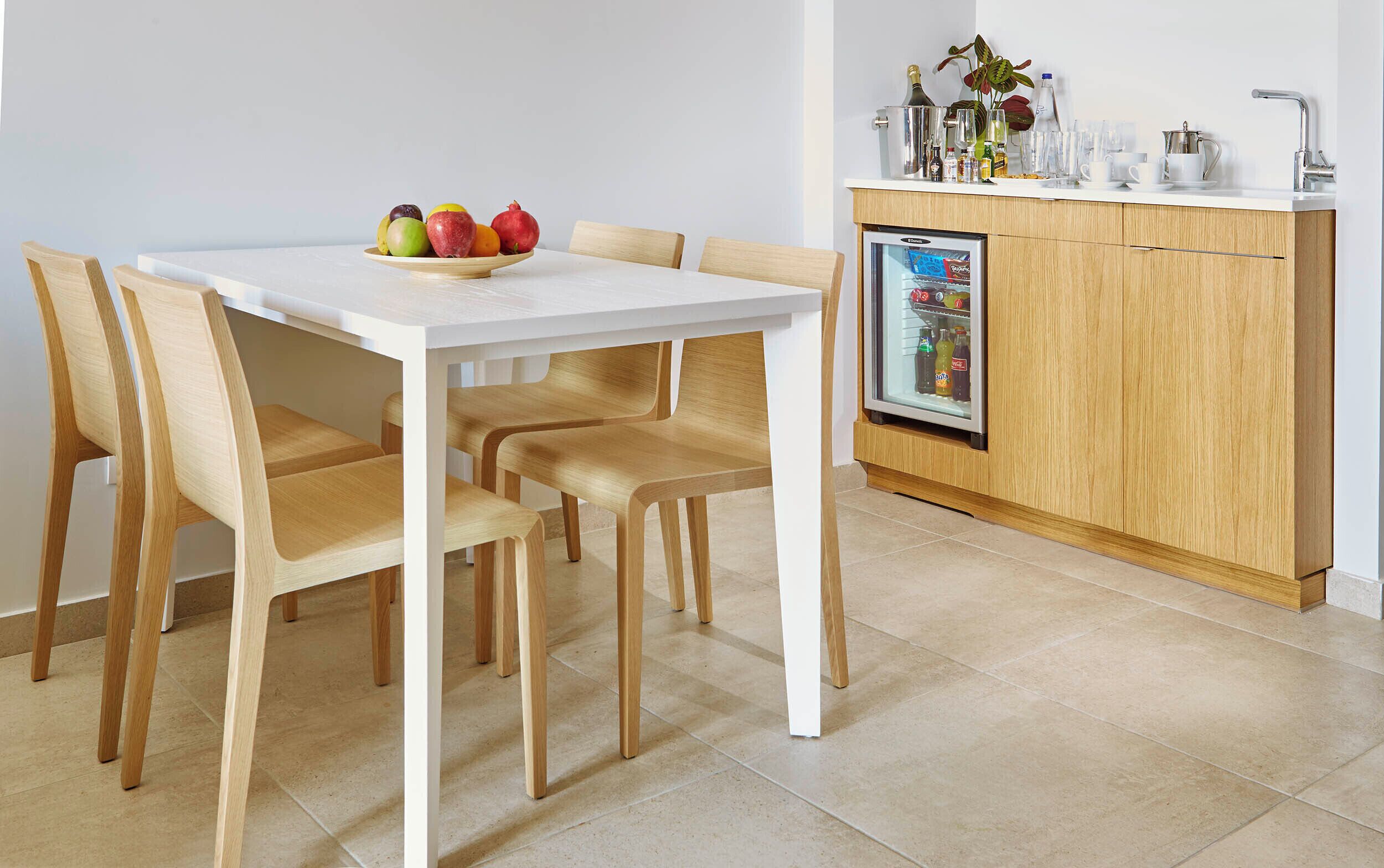 A modern kitchen with a white table and wooden chairs, a refrigerator with a beverage display, and a countertop with various items.