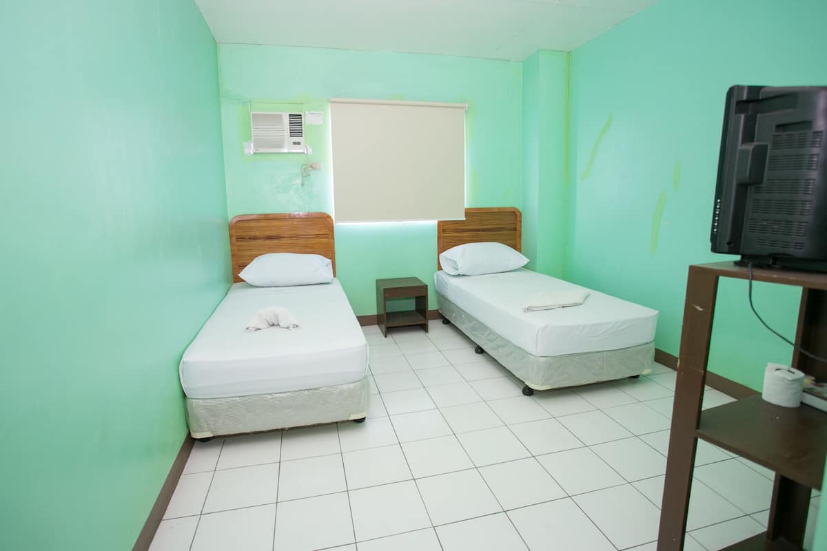 A room with two single beds, a small bedside table, a television on a stand, and a window with blinds.