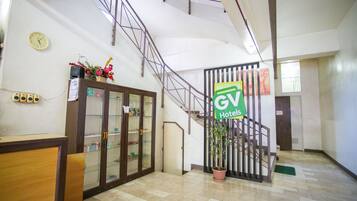 Zona de recepción de hotel con mostrador, vitrina de cristal, escalera y planta en maceta.