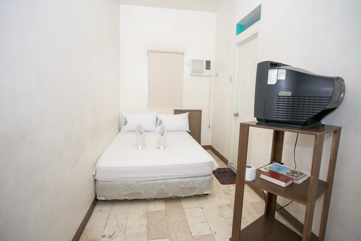 Una habitación pequeña y limpia con una cama, un televisor sobre un mueble de madera y una puerta que da a otra habitación.