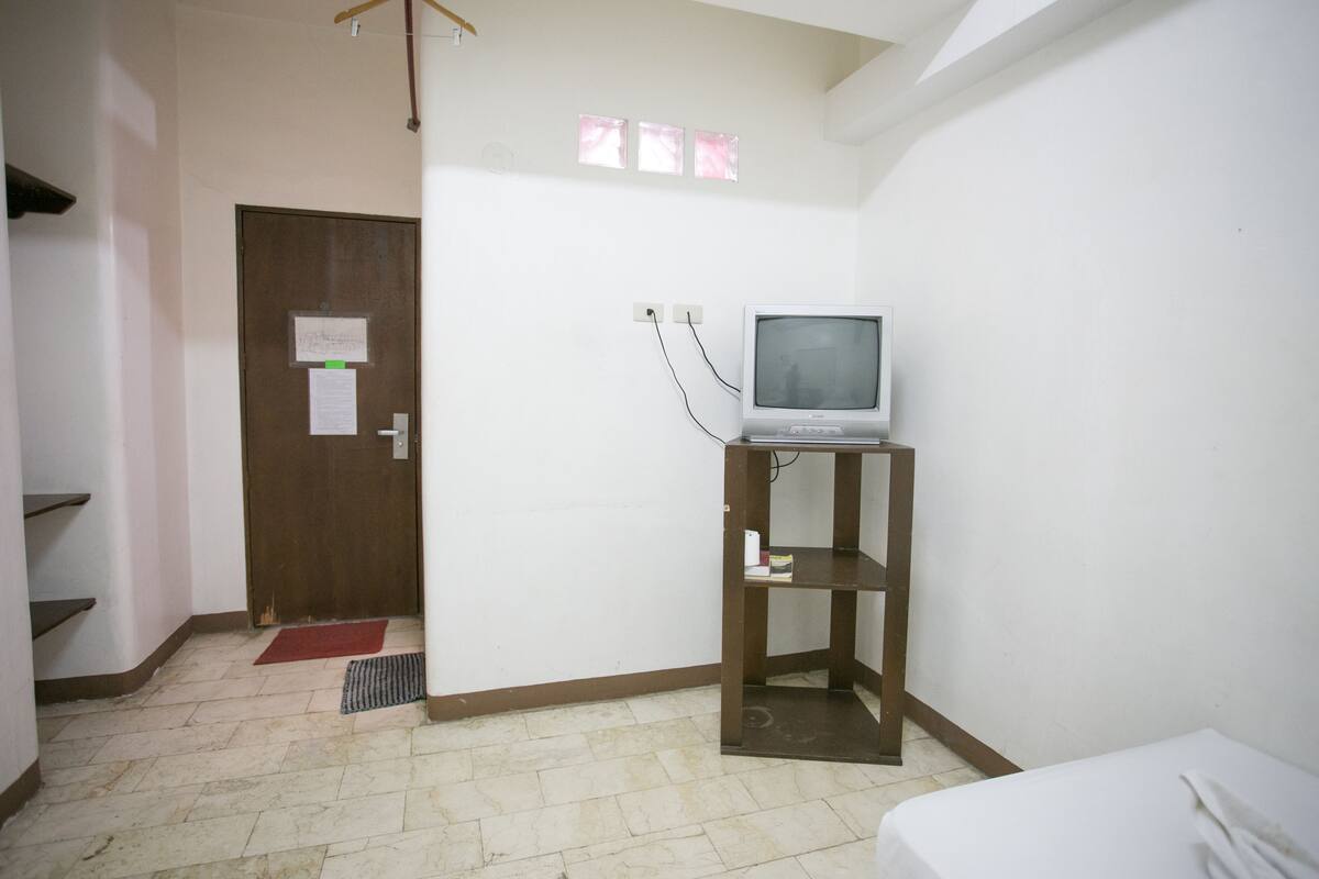 A small, clean room with a single bed, a wooden shelf with a television, a door with a notice, and a tiled floor.