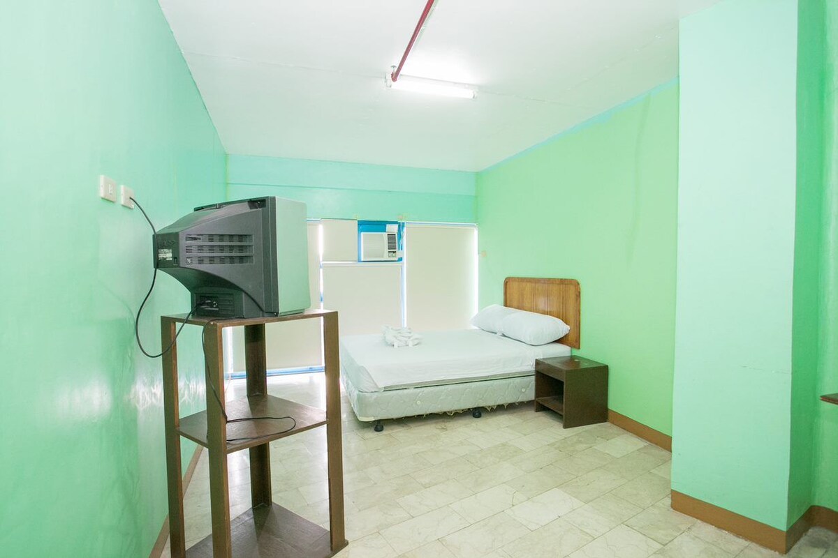 Una habitación de hotel pequeña y sencilla, con cama, mesita de noche y televisor sobre un soporte.