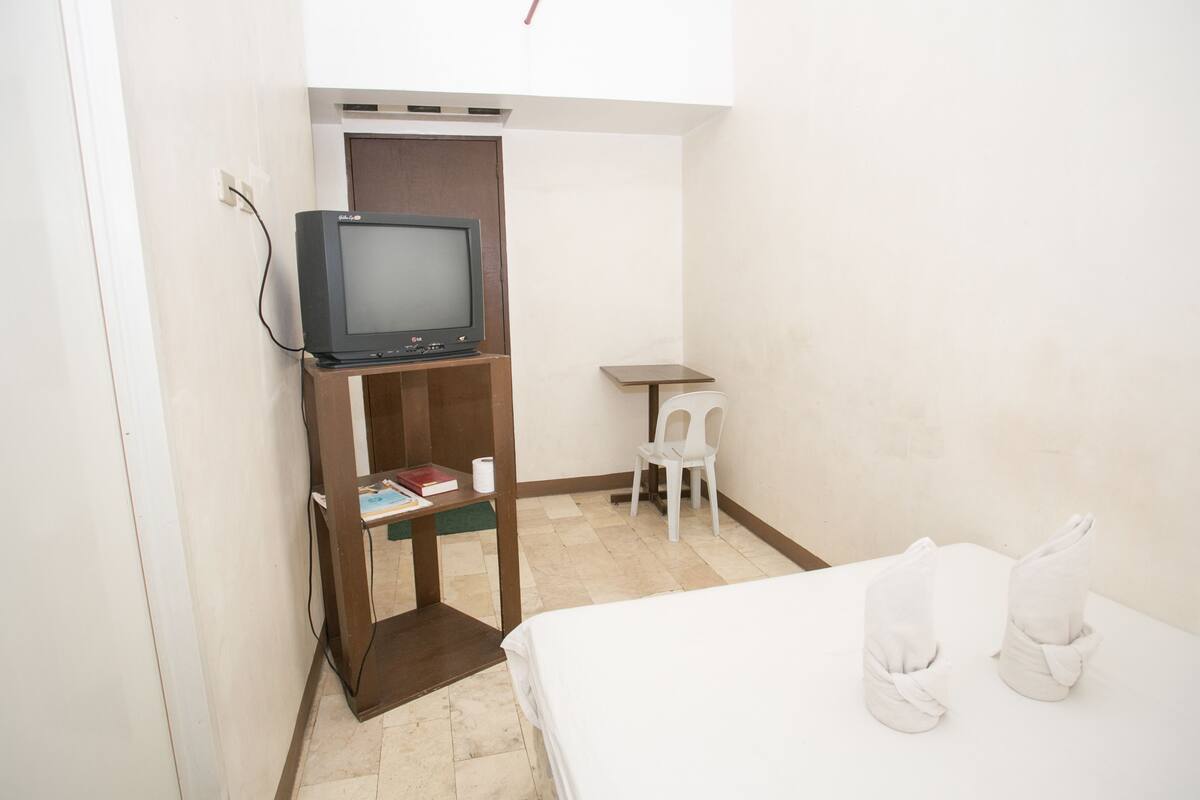 A small room with a bed, a television on a wooden stand, a small table, and a white chair.