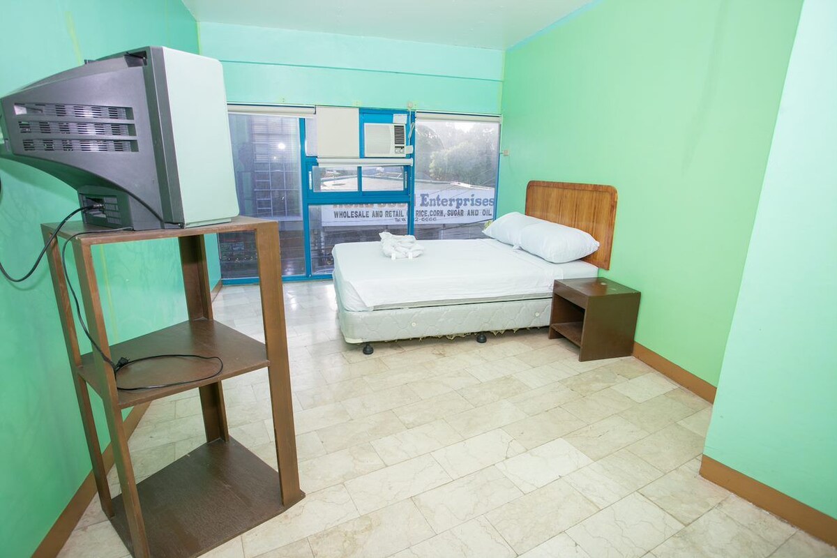 Hotelkamer met een bed, een houten nachtkastje, een televisie op een standaard en een raam met uitzicht op een gebouw.