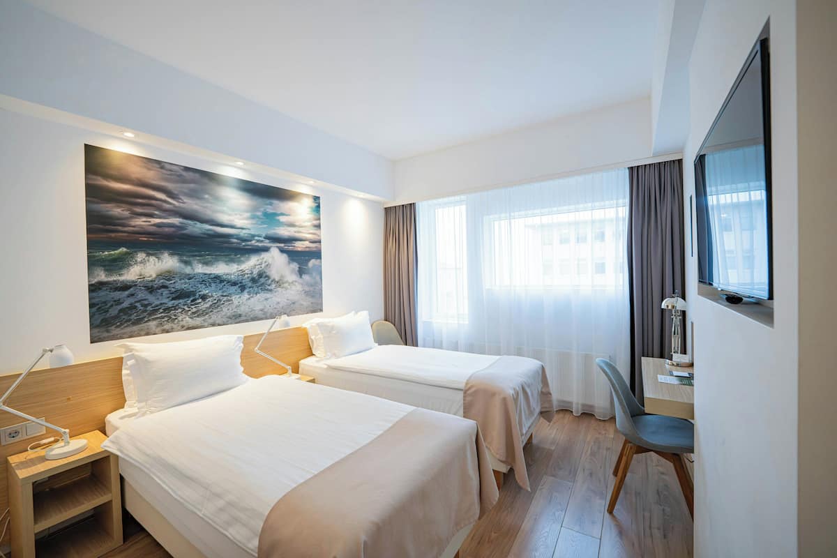 Una habitación de hotel con dos camas, un escritorio, una silla, un televisor y un cuadro grande de un mar tormentoso.