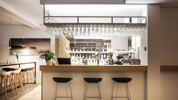 Un área de bar moderna con mostrador de madera, taburetes y una variedad de botellas y vasos.