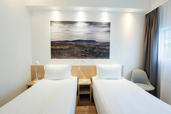 Habitación de hotel con dos camas individuales, una mesita de noche con lámpara, una silla y un cuadro grande con un paisaje.