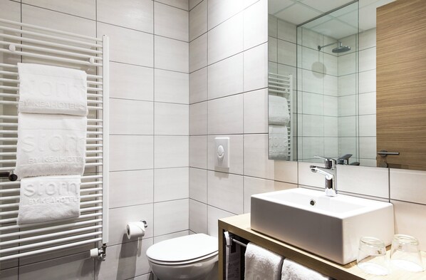 Un baño moderno con pared de azulejos blancos, un inodoro blanco, un lavamanos con espejo y un perchero con toallas.