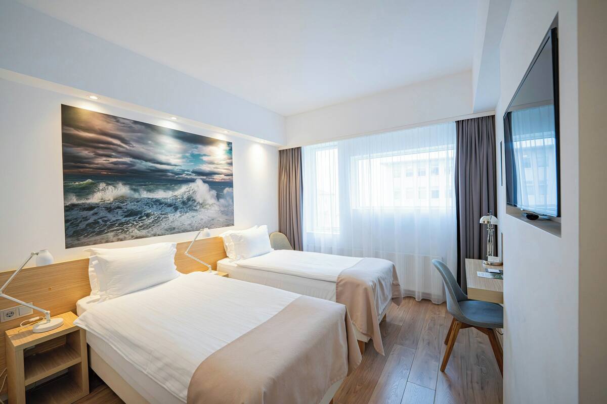 Une chambre d’hôtel avec deux lits, un bureau, une chaise, une télévision et un grand tableau représentant une mer déchaînée.