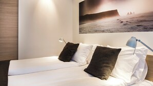 Une chambre à coucher avec un lit, des oreillers, une table de chevet, une lampe et un grand tableau représentant une scène de plage.