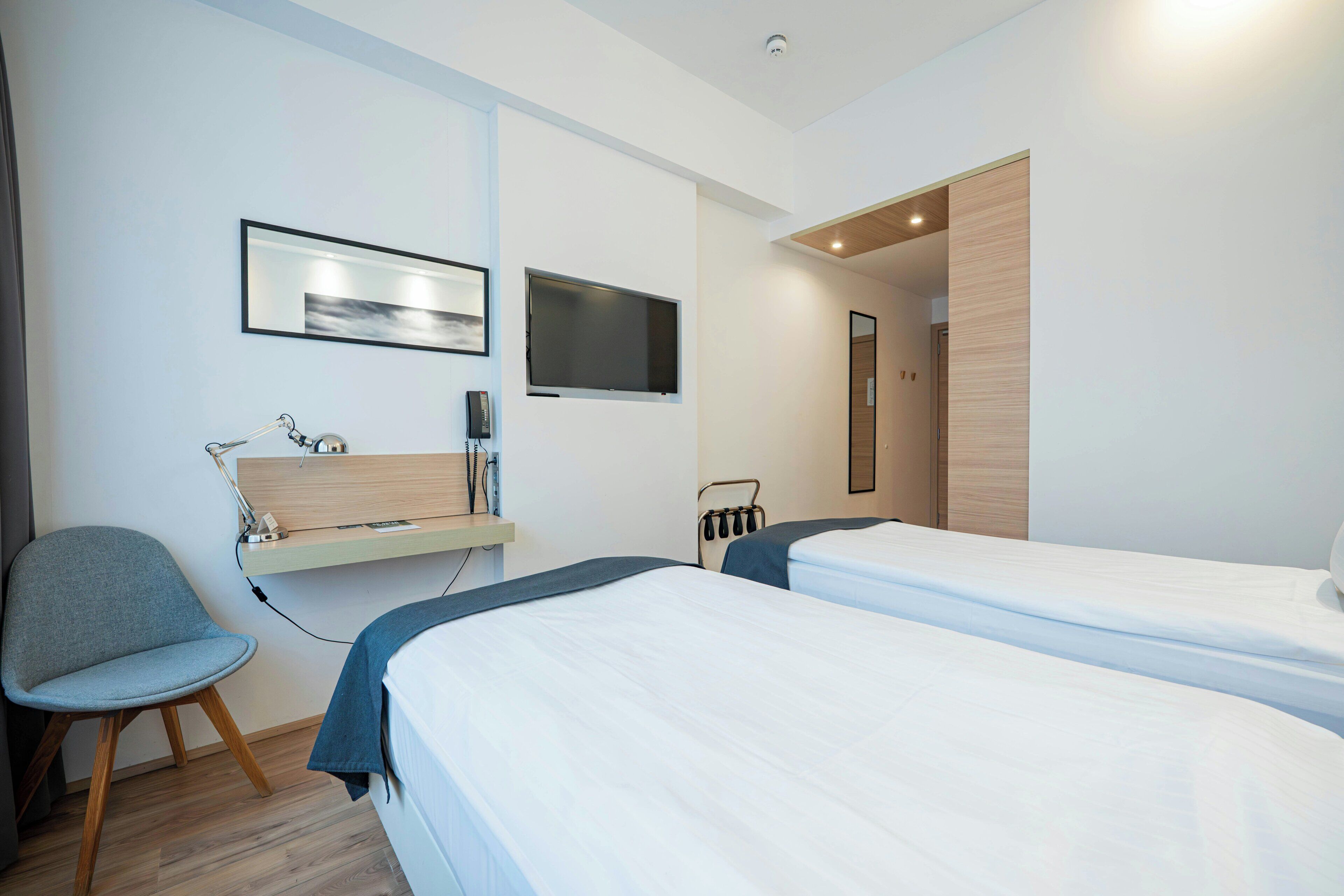 Une chambre d’hôtel moderne équipée de deux lits, d’un bureau avec un ordinateur, d’une chaise et d’un téléviseur fixé au mur.