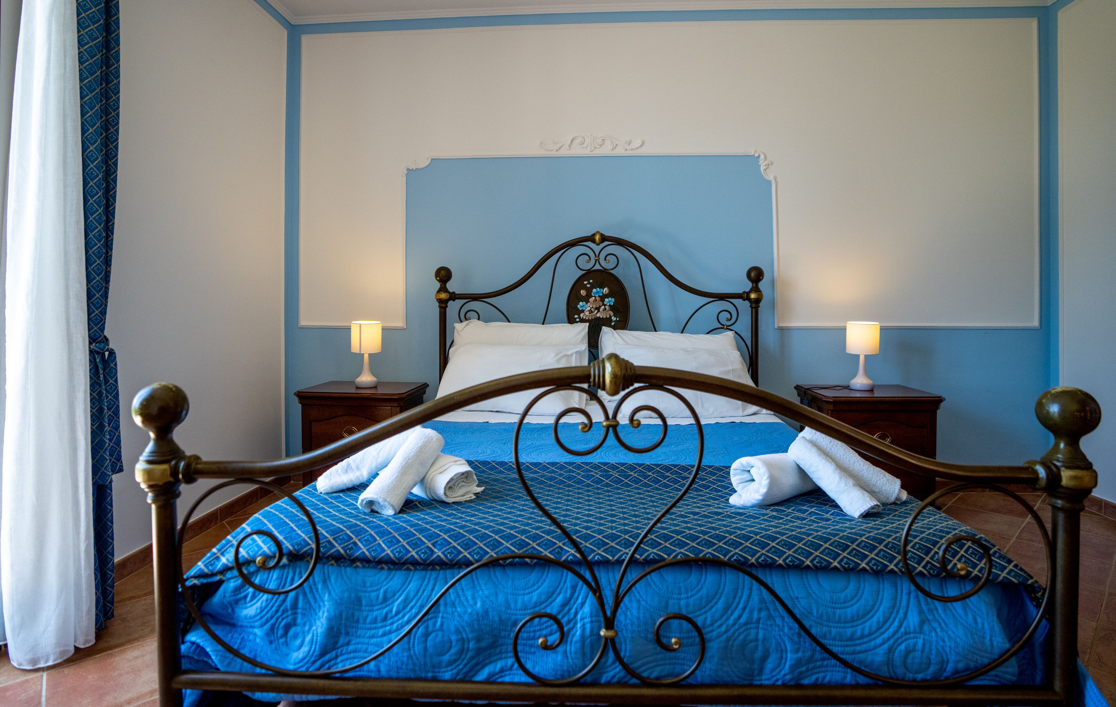 Ein ordentlich gemachtes Bett mit blauer Bettwäsche und weißen Handtüchern, zwei Nachttische mit Lampen und eine blaue Wand mit dekorativer Decke.