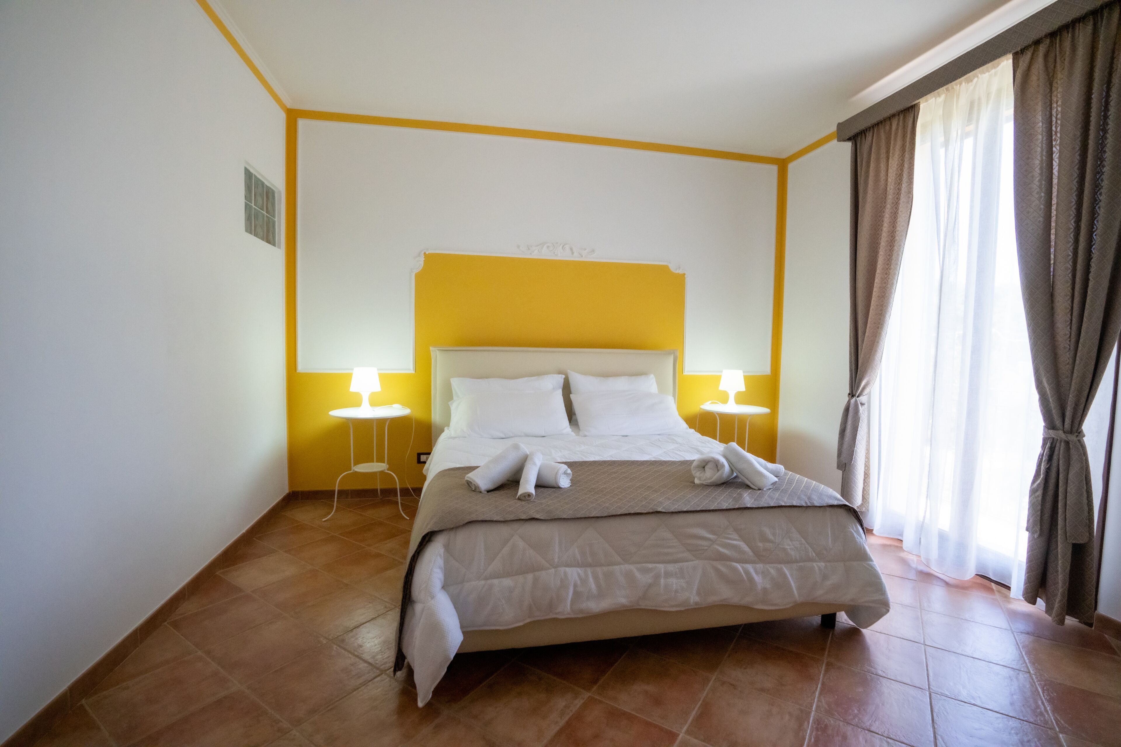 A hotel room with a bed, two bedside tables, a window with curtains, and a yellow accent wall.