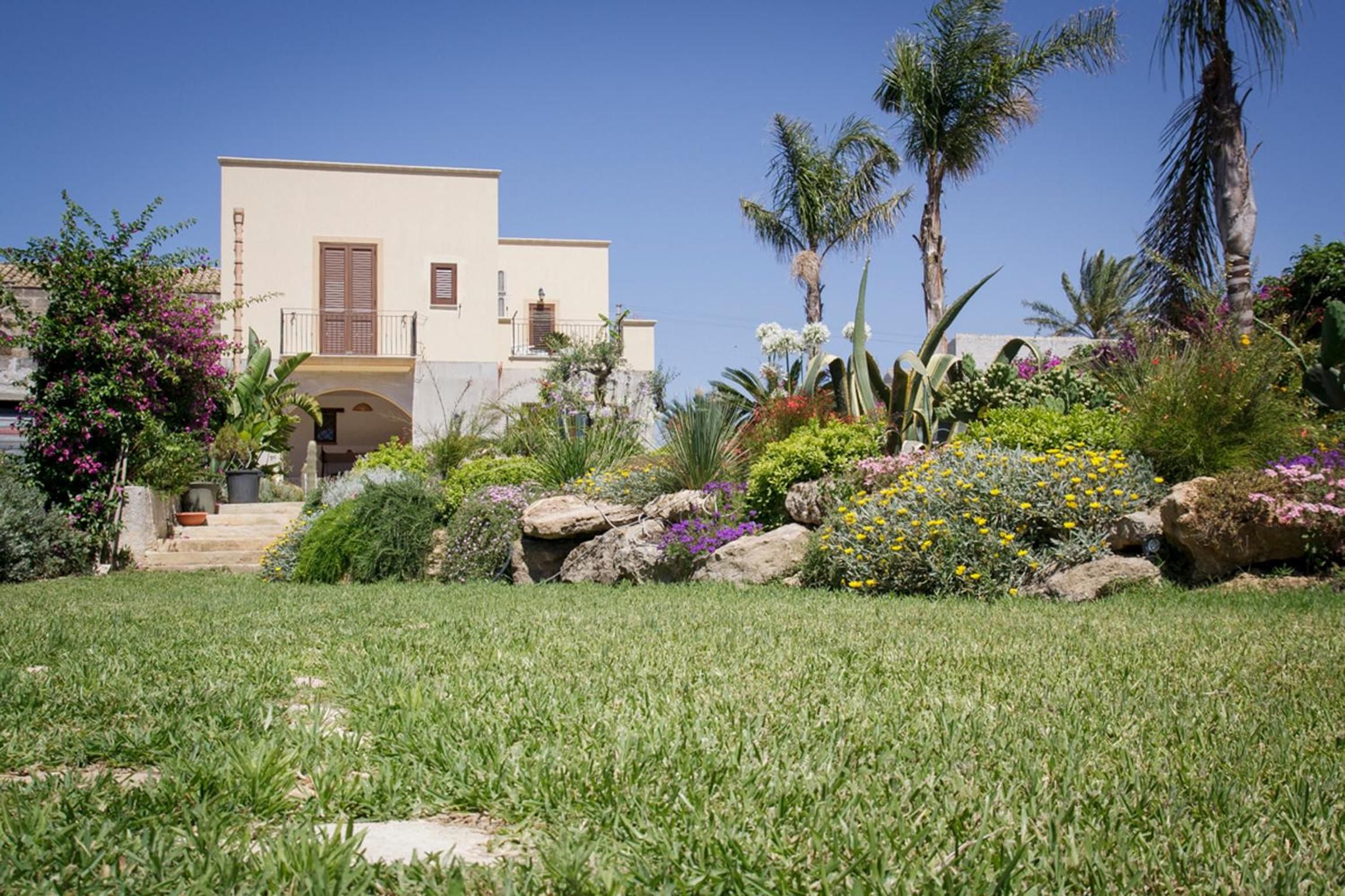 A two-story building with a flat roof, a balcony, and a garden with various plants and flowers.