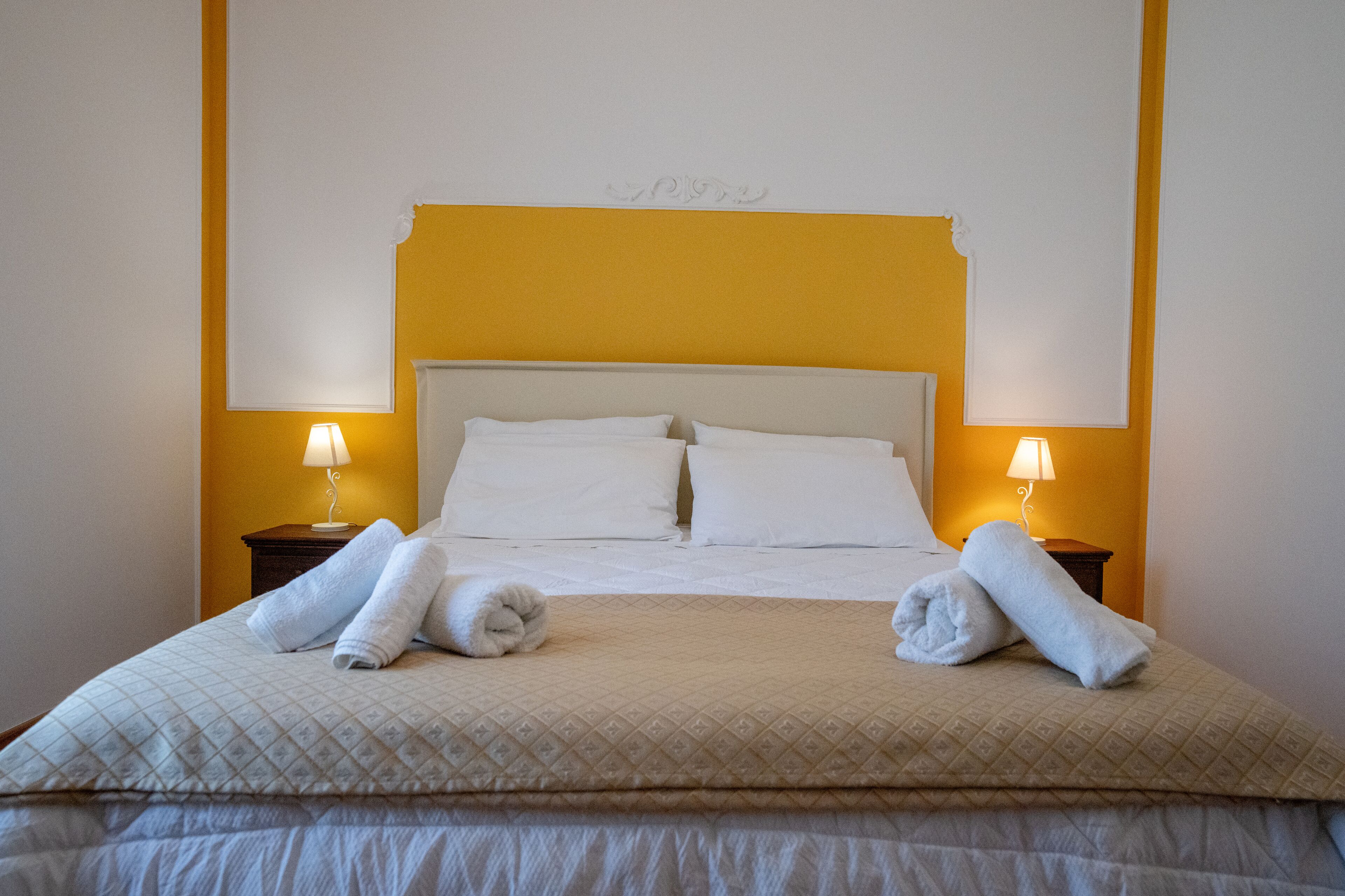 A neatly made bed with a quilted cover, two pillows, and two rolled towels on the bed. There are two bedside tables with lamps on either side of the bed.