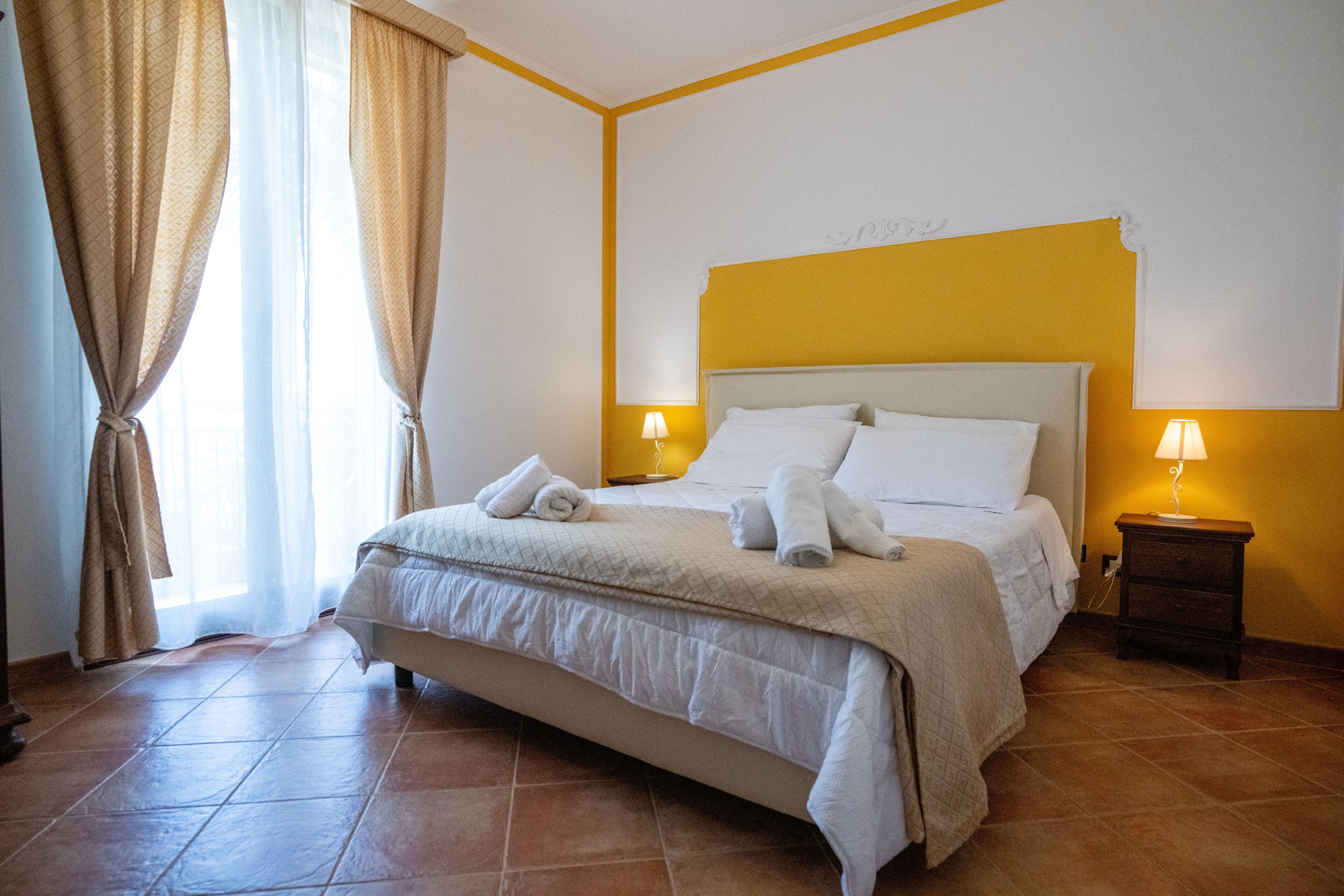 A neatly made bed with white linens and towels, a bedside table with a lamp, and a window with curtains.