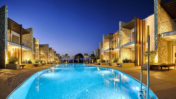Piscina con fachadas de piedra e iluminación cálida, rodeada de mobiliario exterior y palmeras.