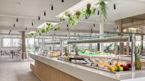 Una barra de desayuno moderna con diversas frutas y bebidas, rodeada de zonas de estar y amplias ventanas.