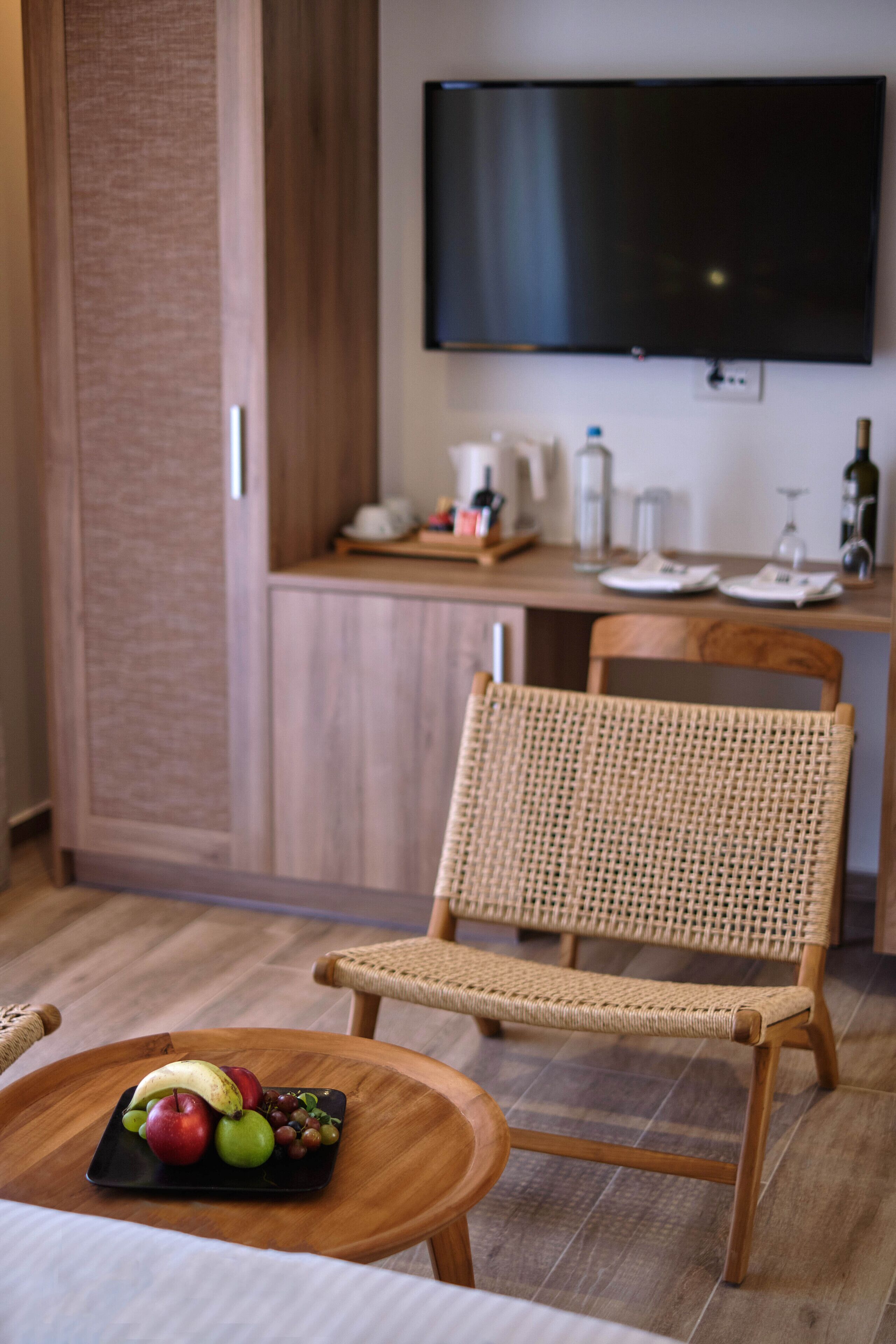 A modern room with a wooden chair, a small table with fruit, a flat-screen TV, and a kitchen area with a coffee maker and wine bottle.
