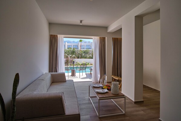 A modern living room with a sofa, a dining table with plates and fruit, and a view of a pool and mountains. - Utopia Blu (Kos)