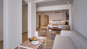 Una sala moderna con un sofá, una mesa de centro de madera con frutas y utensilios, una zona de cocina con una mesa de comedor y sillas, y una pequeña área de descanso con taburetes de mimbre.
