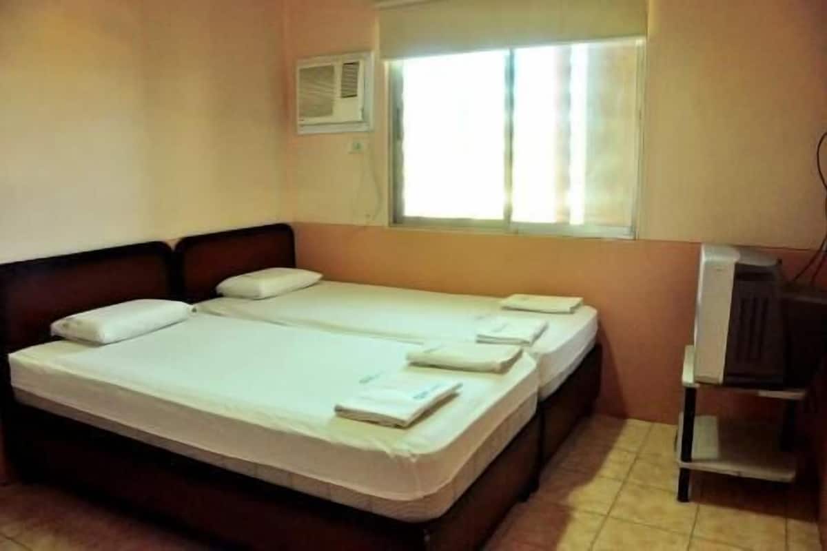 A room with two beds, a window with blinds, a small desk, and a wall-mounted air conditioner.