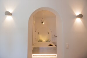 A minimalist hotel room with an arched doorway leading to a bed with two pillows, a hat on the bed, and a small table.