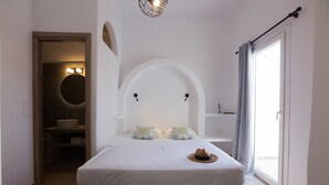 A bedroom with a bed, a hat on the bed, a circular light fixture above the bed, a window with curtains, and a bathroom visible through an archway. - Akti Kastraki Beach (Naxos)
