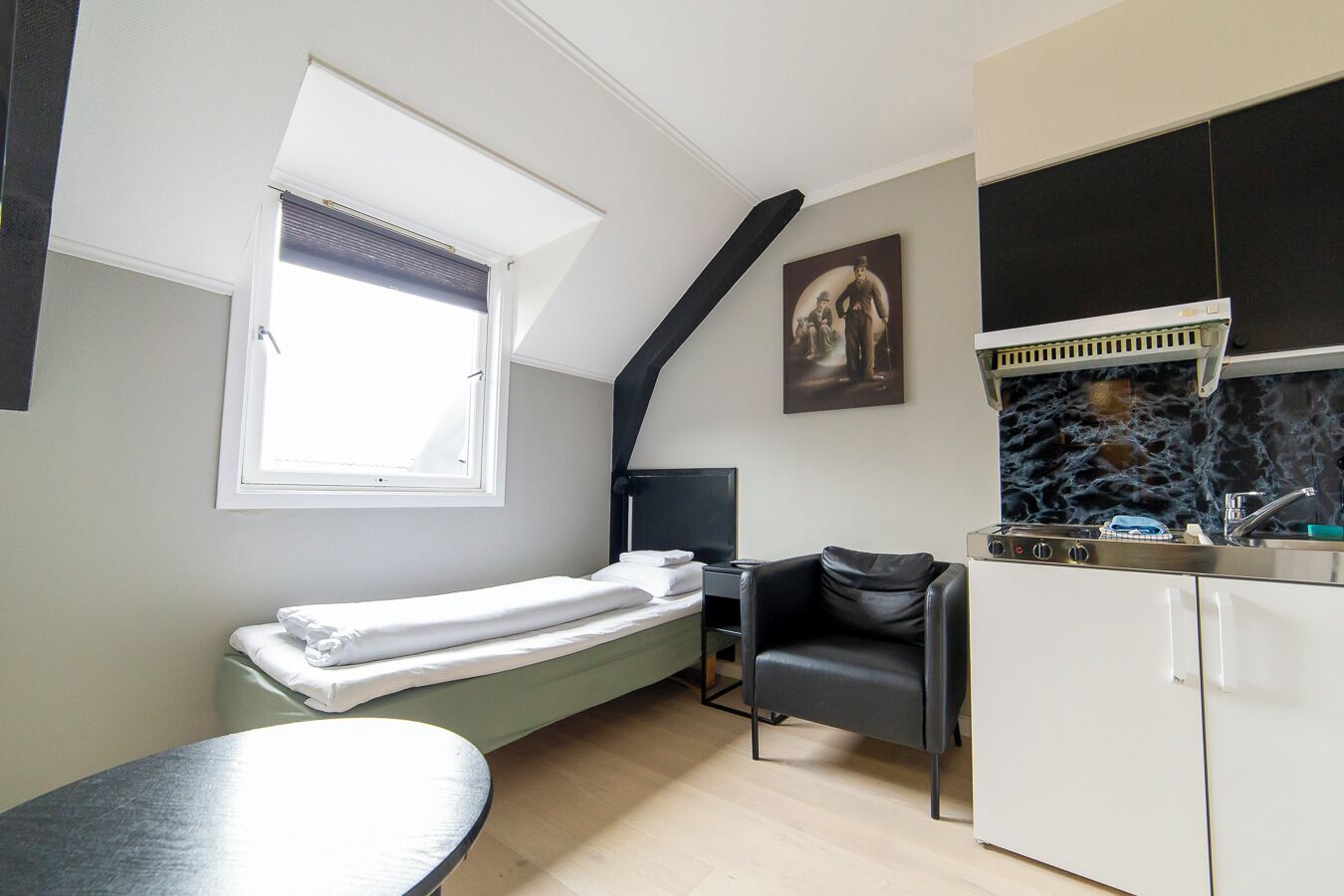 A compact room with a bed, a small table, a chair, a kitchenette, and a window with blinds.