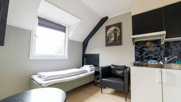 A compact room with a bed, a small table, a chair, a kitchenette, and a window with blinds.